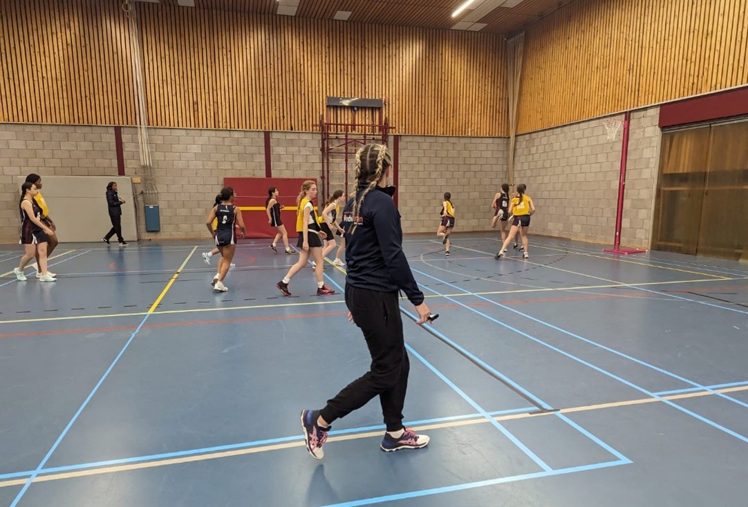 A blonde female with her back to the camera, umpiring a netball match