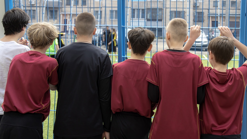 Boys team in burgundy jerseys watching football training behind a fence
